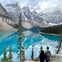Two people stand overlooking a turquoise lake surrounded by tall pine trees, with snow-capped mountains in the background.