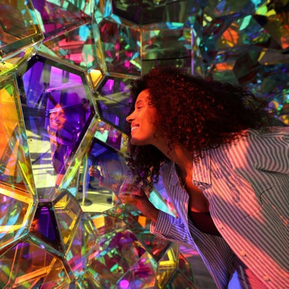 A person in a striped shirt interacts with a colorful, reflective, geometric structure under vibrant lighting.