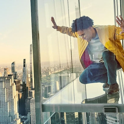 A person crouches on a glass balcony high above a cityscape, surrounded by skyscrapers and a clear sky.