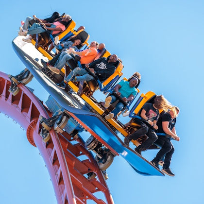 People riding a rollercoaster on a steep incline under a clear blue sky, showing excitement and various expressions.