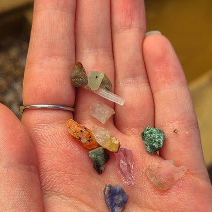 A hand holding various small, colorful crystals and minerals. A silver ring is visible on the person's finger.