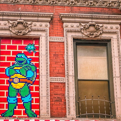 Brick building with ornate window frames. Left frame has a "Teenage Mutant Ninja Turtle" mosaic; right window has a metal grate.