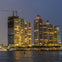 High-rise buildings with illuminated windows at dusk, one under construction, with a waterfront in the foreground.