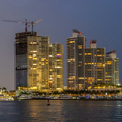 High-rise buildings with illuminated windows at dusk, one under construction, with a waterfront in the foreground.
