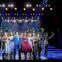 A group of performers in formal attire stand on a lit stage holding hats, with theatrical lighting and set design in the background.