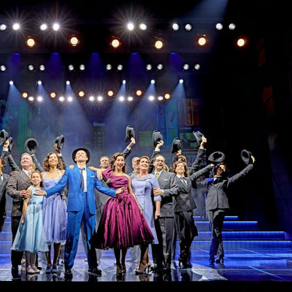 A group of performers in formal attire stand on a lit stage holding hats, with theatrical lighting and set design in the background.