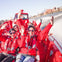 A group of people wearing red jackets and life vests enjoy a boat ride, smiling and waving, with a cityscape in the background.