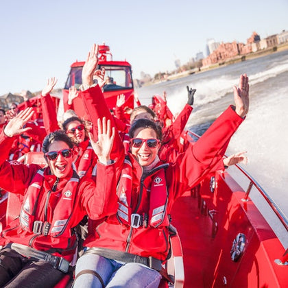 A group of people wearing red jackets and life vests enjoy a boat ride, smiling and waving, with a cityscape in the background.