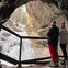 Two people in winter clothing stand in a cave, looking out at a snowy landscape with a frozen waterfall.
