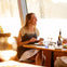 A woman smiles while dining at a restaurant table with food, drinks, and sunlight streaming through the window.