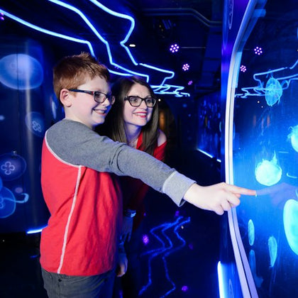 Two people smiling and pointing at a glowing interactive display with blue neon lights and jellyfish graphics in a dark room.