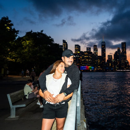 A couple embraces near a waterfront rail, with people seated on benches and a city skyline glowing in the twilight.