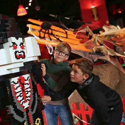 Two boys interact with a large LEGO vampire figure. Spiders and cobwebs decorate the background in a Halloween-themed setup.