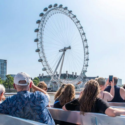 People seated on a boat facing a large Ferris wheel, with a cityscape visible in the background under a clear sky.