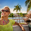 A woman in sunglasses and a neon top rides in an open vehicle, with palm trees and a house near water in the background.