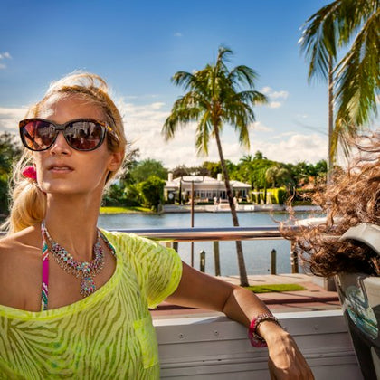 A woman in sunglasses and a neon top rides in an open vehicle, with palm trees and a house near water in the background.