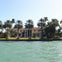 A waterfront mansion with a red tile roof, surrounded by palm trees, with a green lawn and water in the foreground.