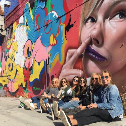 A group of women sitting on the sidewalk in front of a colorful graffiti wall featuring a large face mural.