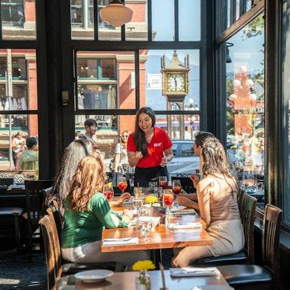 A group of people seated at a restaurant table being served by a waitress. Large windows and a street view are in the background.