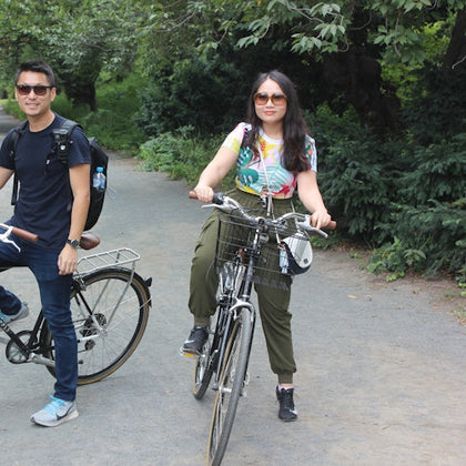 Two people on a path with bicycles, surrounded by greenery and trees.