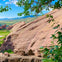 Large reddish rock formations surrounded by greenery under a blue sky with scattered clouds, framed by leafy branches.