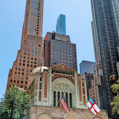 A historic building with an arched facade and two flags in front, surrounded by tall skyscrapers on a clear day.