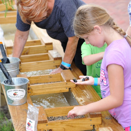 A girl examines rocks in her hand while participating in a gem mining activity. An adult assists in the background.
