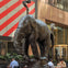 A bronze elephant sculpture with an unusual shape in a public plaza; people are seated nearby at outdoor tables.