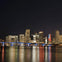 A brightly lit city skyline at night with a bridge illuminated in blue lights and its reflection visible on the water.