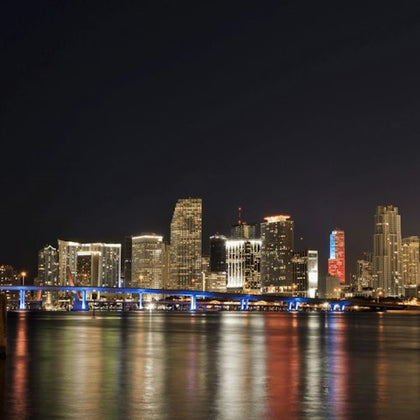 A brightly lit city skyline at night with a bridge illuminated in blue lights and its reflection visible on the water.