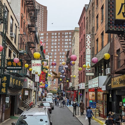 A bustling urban street lined with shops and restaurants, decorated with colorful lanterns overhead, and people walking.