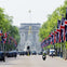 Buckingham Palace with its iconic gates, Union Jack flags lining the street, and traffic including taxis and a motorbike.