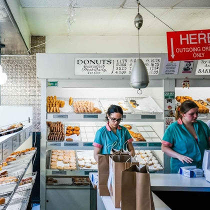 Two employees in a donut shop prepare orders with pastries behind them and filled paper bags on the counter.
