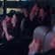A group of people sitting in a theater clapping and smiling during a performance, with dim lighting and blurred background.