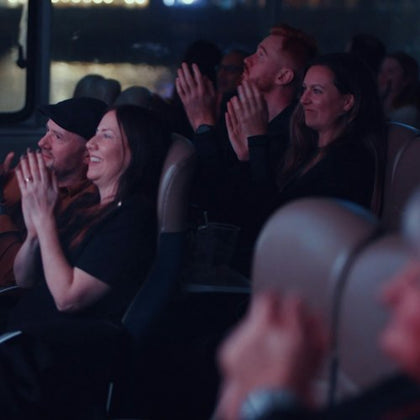 A group of people sitting in a theater clapping and smiling during a performance, with dim lighting and blurred background.