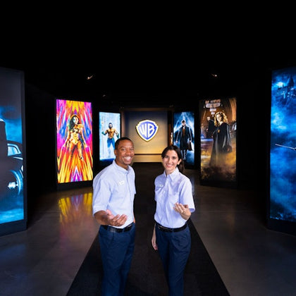 Two people in uniform stand in a hallway with colorful movie-themed panels and a Warner Bros. logo in the background.