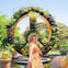 A woman in colorful attire and a hat stands in front of a large floral peace sign in a garden setting.