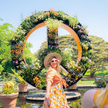 A woman in colorful attire and a hat stands in front of a large floral peace sign in a garden setting.