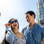 Two smiling people, a woman in sunglasses and a man in a blue shirt, outdoors near a building with "UNIVERSAL" signage.