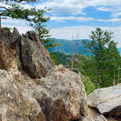 Rocky outcropping surrounded by coniferous trees with a forested mountain in the background under a partly cloudy sky.