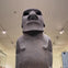 A large stone moai statue displayed indoors, illuminated by ceiling lights, set against a plain background.