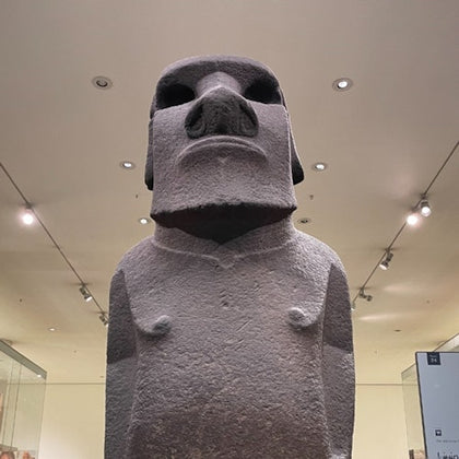 A large stone moai statue displayed indoors, illuminated by ceiling lights, set against a plain background.
