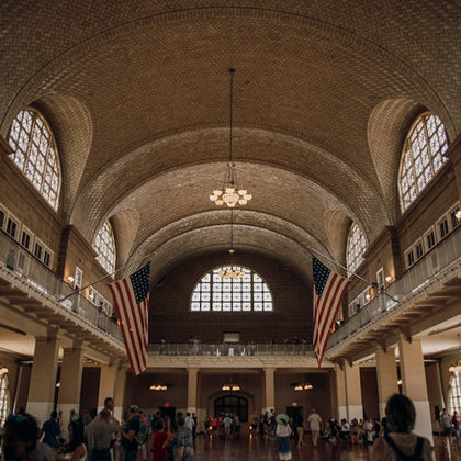 A large hall with an arched ceiling, chandeliers, arched windows, and American flags. People are gathered below, some sitting, some standing.
