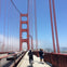 A cyclist and pedestrians on a walkway of the Golden Gate Bridge, with cars on the road and fog in the background.