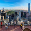 People on a rooftop observation deck, photographing a city skyline with skyscrapers and a central park under a blue sky.