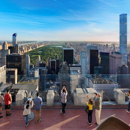 People on a rooftop observation deck, photographing a city skyline with skyscrapers and a central park under a blue sky.