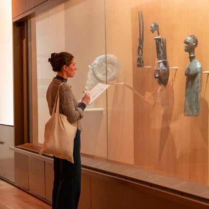 A woman observes sculptures displayed in a museum exhibit, holding a booklet and wearing a shoulder bag.