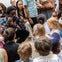 A man animatedly speaks to a group of children and adults gathered around him indoors.
