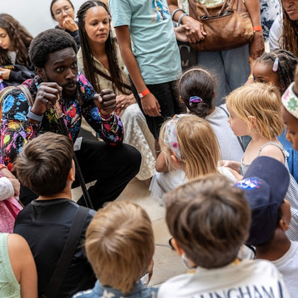 A man animatedly speaks to a group of children and adults gathered around him indoors.