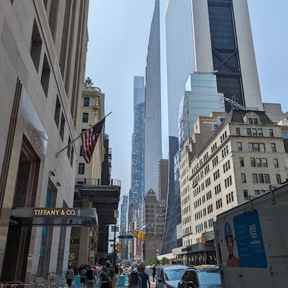 A busy city street with buildings, including Tiffany & Co., and pedestrians under a clear sky with visible skyscrapers.